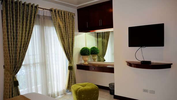 A hotel room with a bed, a green ottoman, a TV on a wall, a mirror above a vanity, and curtains on the windows. - Grand Isabella Residences (Cebu City)