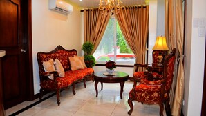 A room with a wooden door, a sofa, a chair, a table, a lamp, and a window with curtains. - Grand Isabella Residences (Cebu City)