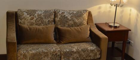 A brown sofa with floral upholstery and a matching cushion, positioned against a wall with a framed picture. A wooden side table with a lamp is to the right.