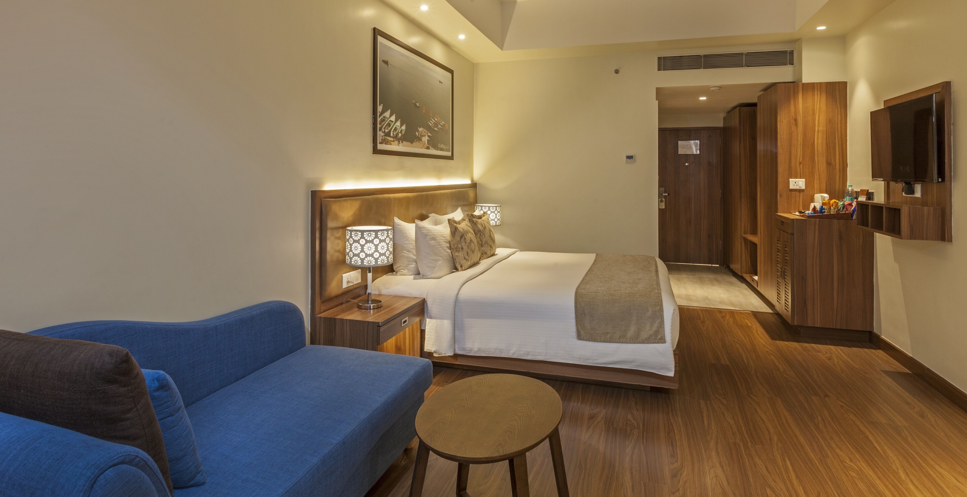 Habitación de hotel con cama, sofá azul, una mesita redonda, televisión y armario de madera.