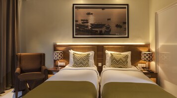 Habitación de hotel con dos camas, una silla, una mesa y un cuadro con barcos en la pared.