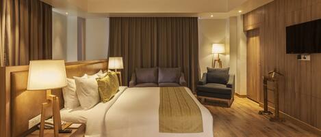 A hotel room with a large bed, two bedside tables with lamps, a chair, a television, and wooden paneling on the walls.