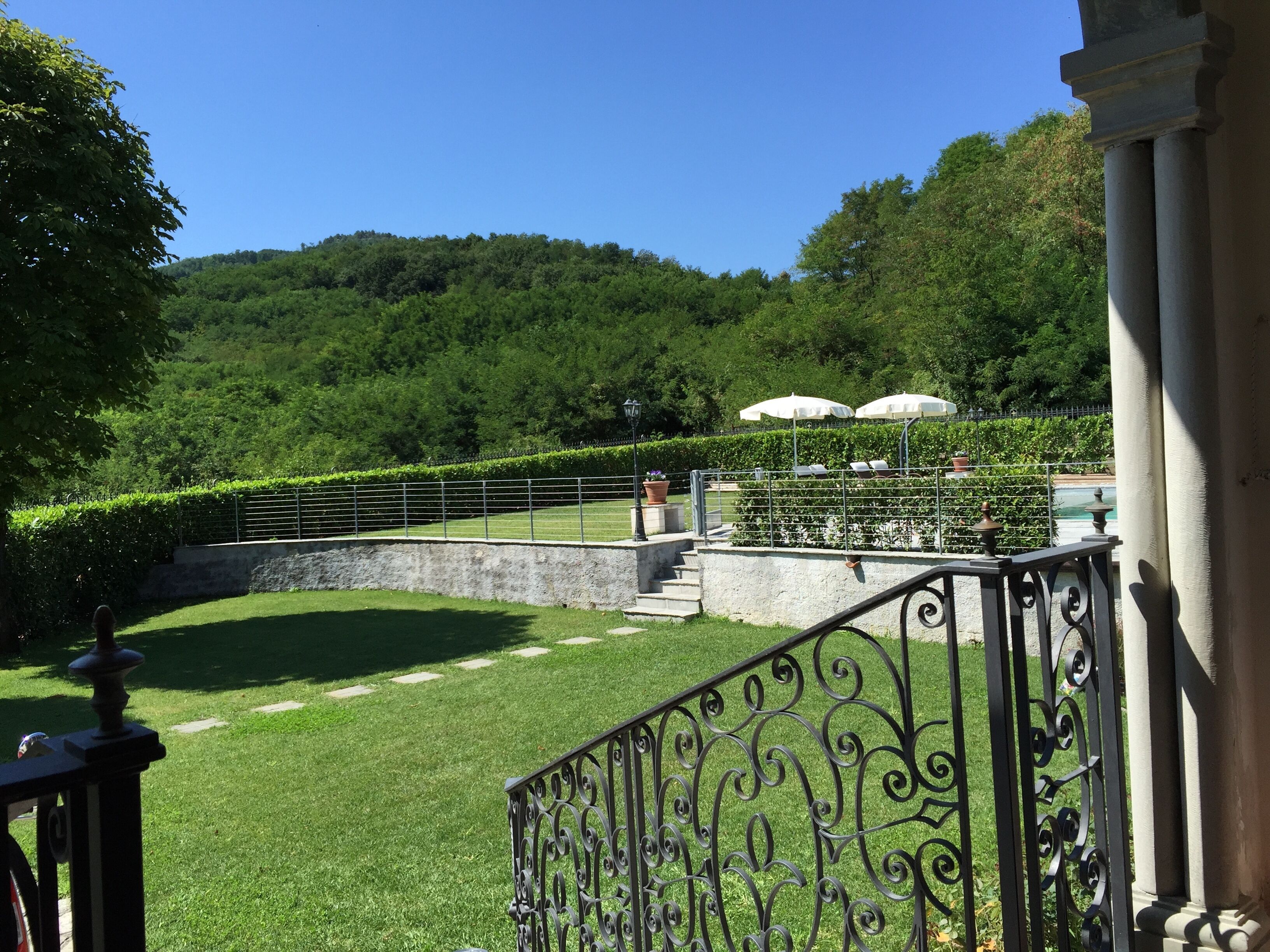 Un jardí paisatjistic amb un mur de pedra, una barana metàl·lica i un camí que condueix a una zona d'estar amb ombrel·les.