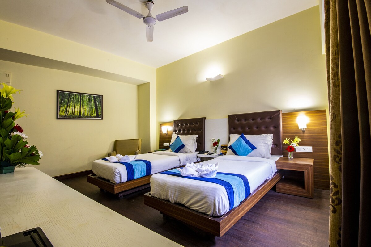 A hotel room with two beds, a ceiling fan, a nightstand with a lamp, and a painting of a forest.