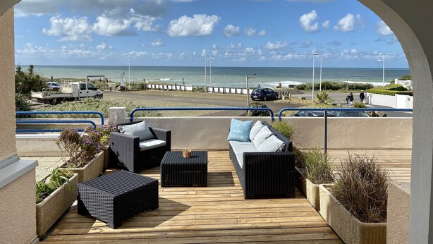 Ein überdachter Außenbereich mit Korbmöbeln und Pflanzgefäßen mit Blick auf Strand und Meer.