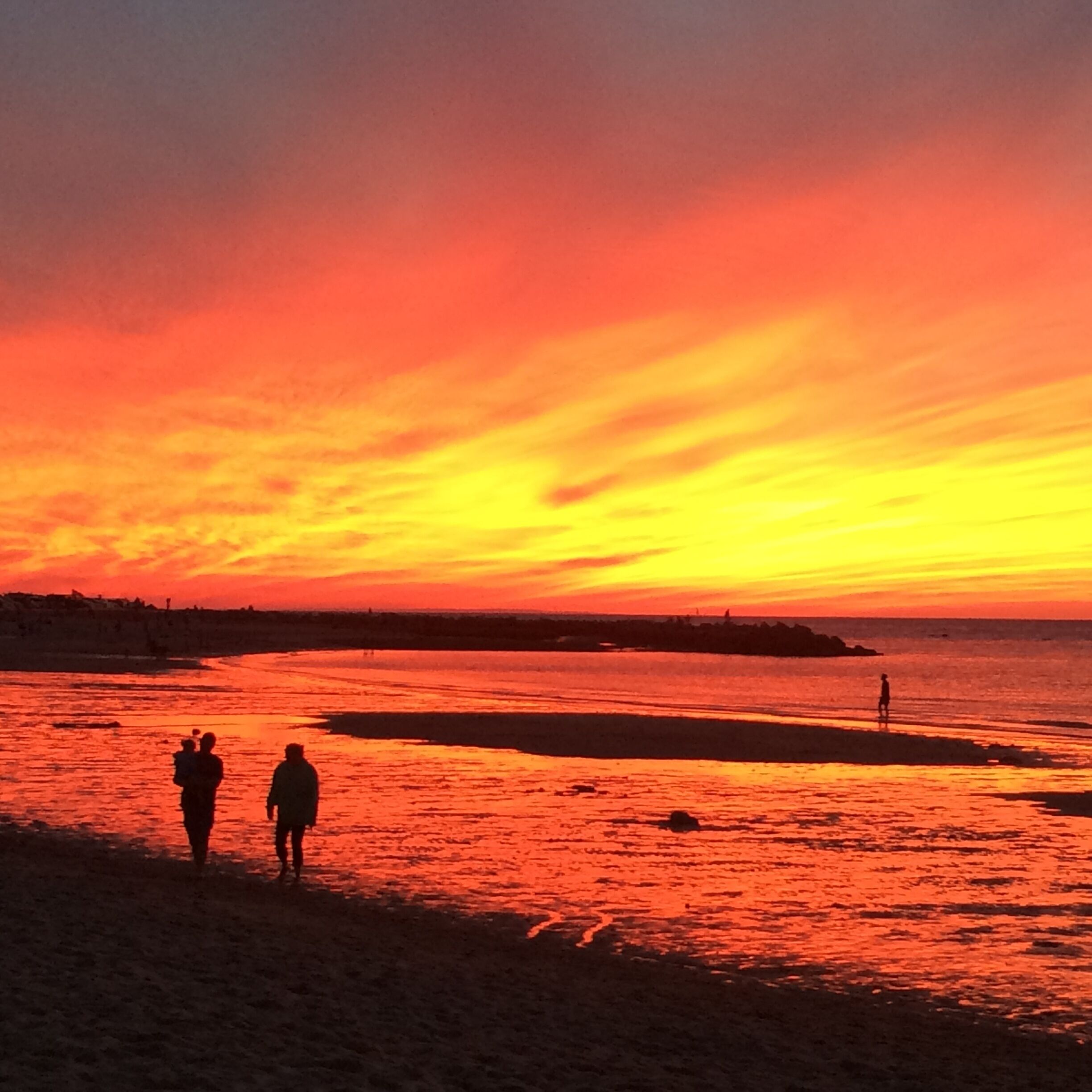 Strandscene i solnedgang med silhuetter af mennesker og en fjerntliggende kystlinje.