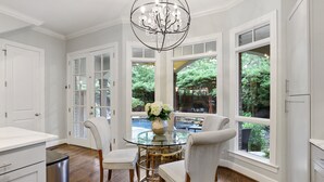 A dining area with a glass table, white chairs, a chandelier, and large windows with a view of greenery. - Modern Tudor with Pool & Backyard Oasis on the M Streets in the Heart of Dallas (Dallas)