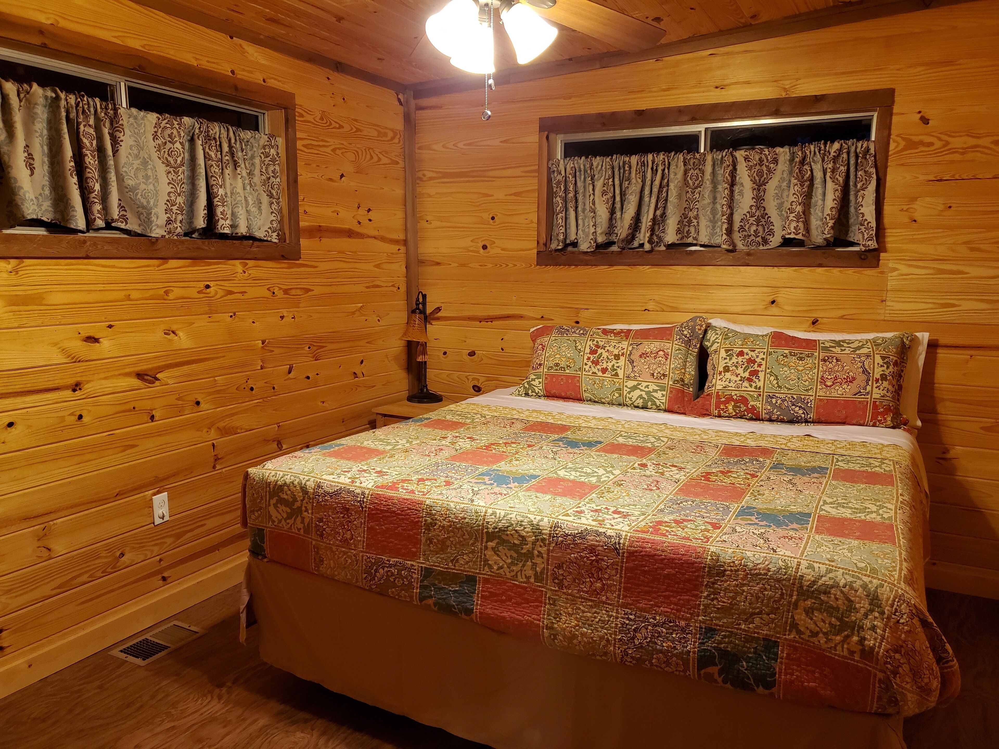 A wooden cabin bedroom with a quilted bedspread, two windows with curtains, a bedside lamp, and a wooden ceiling.