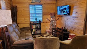 A cozy cabin interior with a wooden interior, a sofa, a bookshelf, a television, and a Christmas decoration. - Treehouse Cabin/ Beach and Lake Access (Pottsboro)