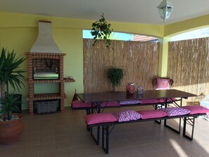 Eine Terrasse im Freien mit einem Backofen aus Ziegeln, einem Tisch mit Bänken, Topfpflanzen und einem Bambus Sichtschutz.