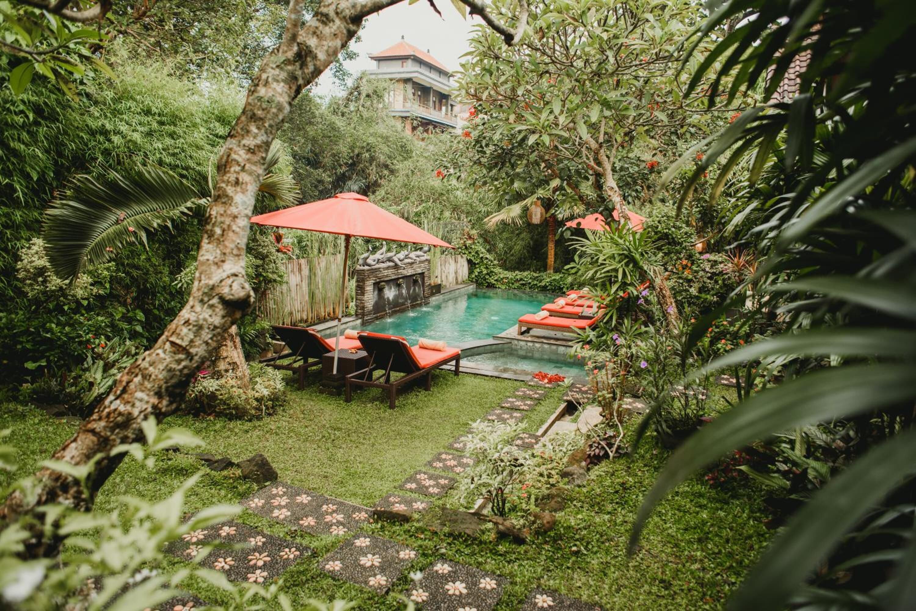 A lush garden with a swimming pool, red umbrellas, and lounge chairs.