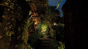 A narrow, stone-paved pathway leading to a wooden structure with a sloped roof, surrounded by lush greenery and trees. - Desak Putu Putra Hidden (Ubud)