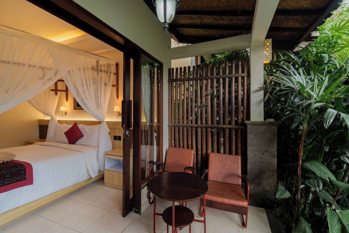 A bedroom with a bed, bedside table, and a small table with chairs. There is a wooden partition with plants behind it.