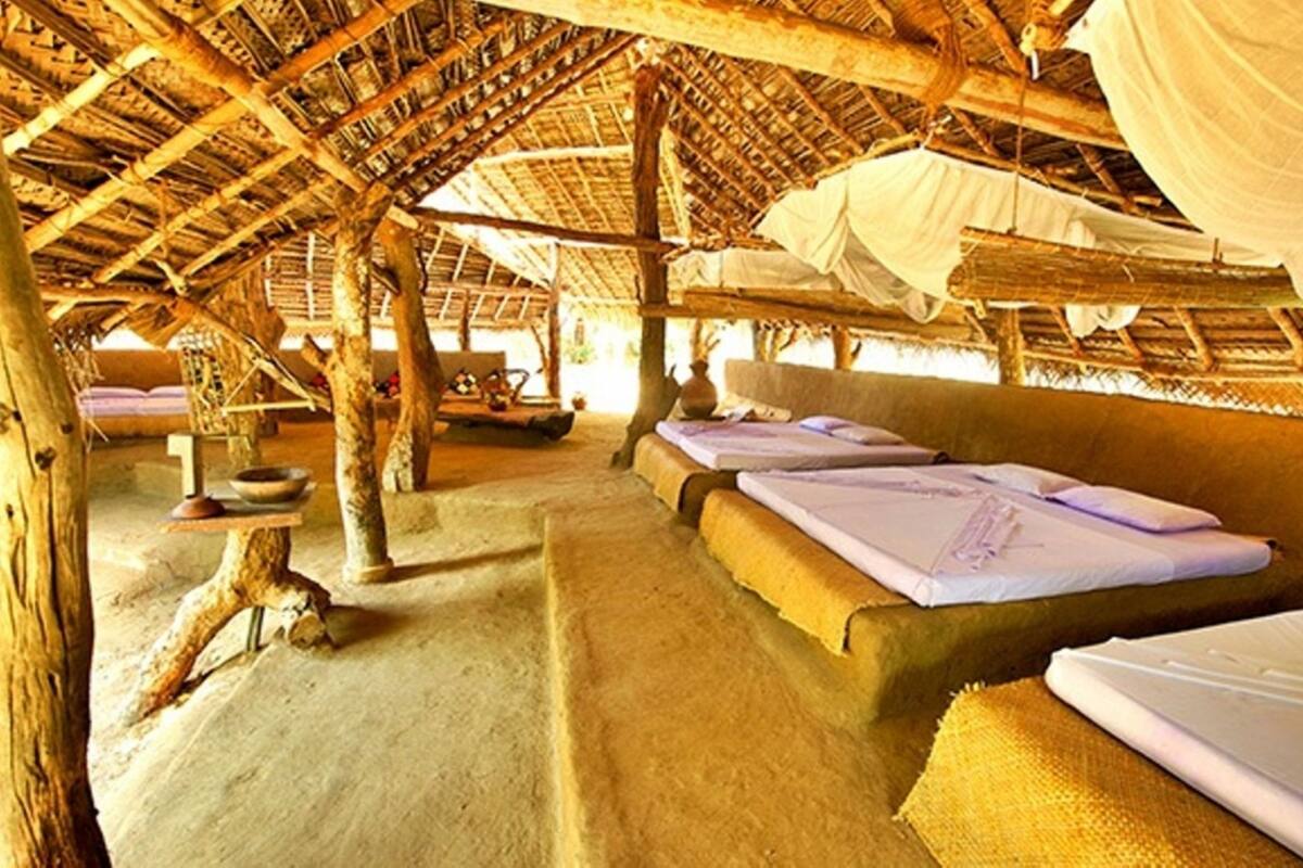 A rustic thatched-roof hut with two beds, a bench, and a small table with a bowl.