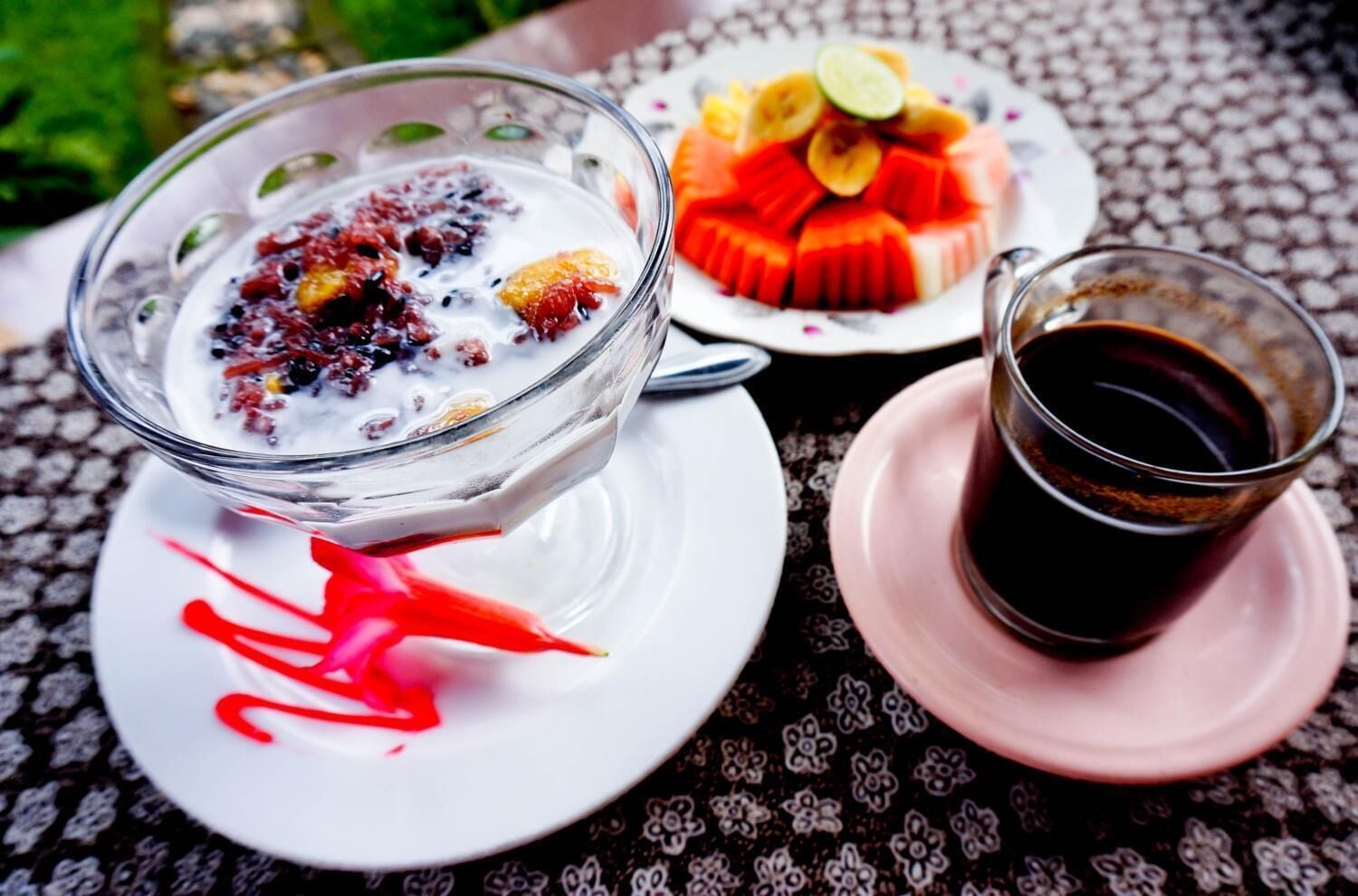 A dessert bowl with fruit and a glass of dark beverage on a patterned tablecloth.