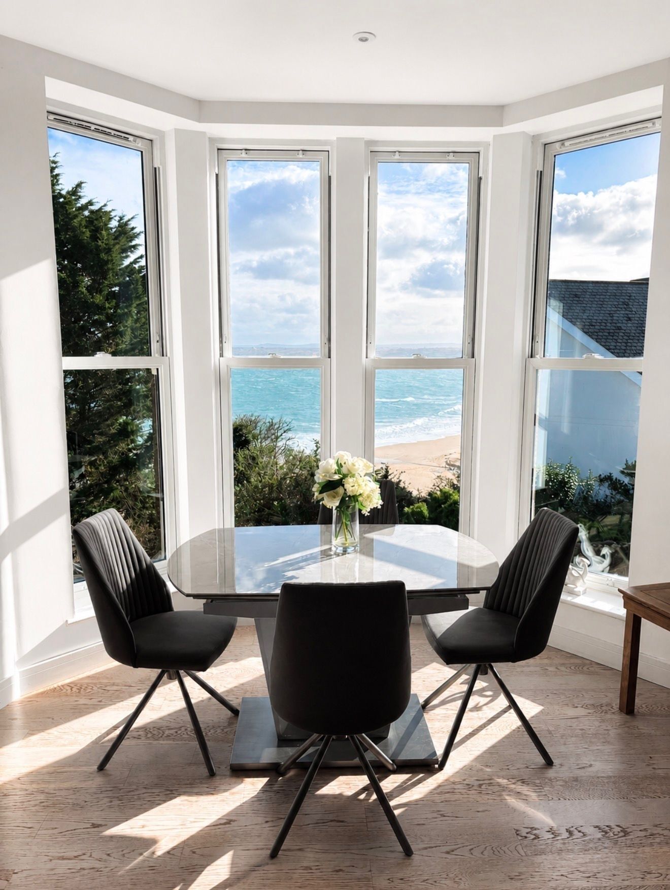 A corner room with a bay window overlooking the sea, a round glass table with two chairs, and a vase with flowers.