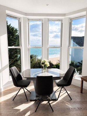 A corner room with a bay window overlooking the sea, a round glass table with two chairs, and a vase with flowers.