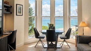 Ein modernes Wohnzimmer mit Erkerfenster mit Meerblick, einem Esstisch mit Stühlen, einem Sofa und einer Lampe.