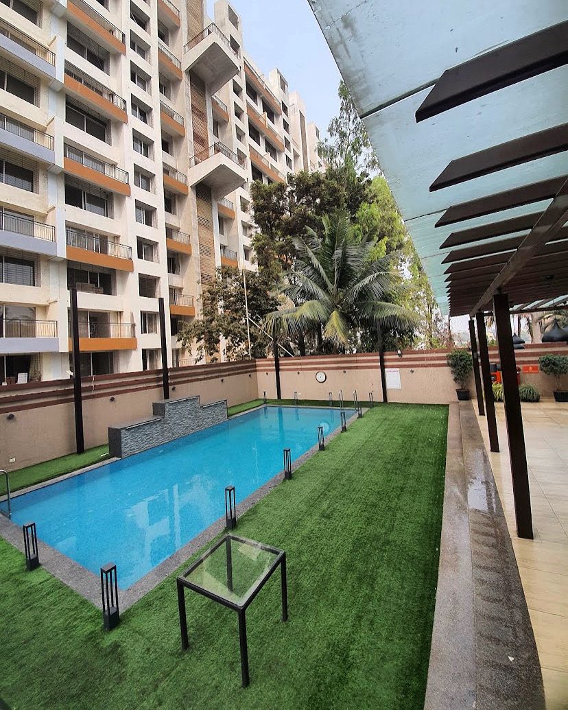 A swimming pool with a grassy area, a covered seating area, and a multi-story building in the background.