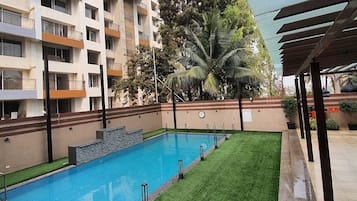 A swimming pool with a grassy area, a covered seating area, and a multi-story building in the background.