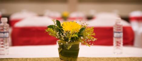 A glass vase with a yellow flower arrangement on a table, surrounded by water bottles and red bedding in the background.