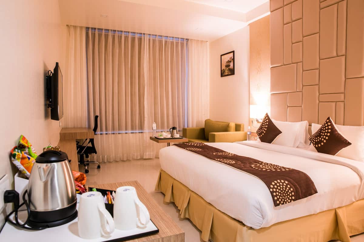 A hotel room with a bed, a desk with a kettle and cups, a chair, a small table with a bottle, and a curtain.
