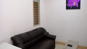 A room with a brown leather sofa, a small table with a remote control, a window with blinds, and a framed picture of a purple flower on the wall. - Bansuannamdee (Pluak Daeng)