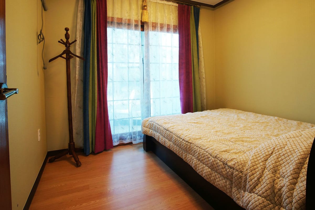 A bedroom with a wooden floor, a bed with a quilted cover, a wooden door, and a window with colorful curtains.