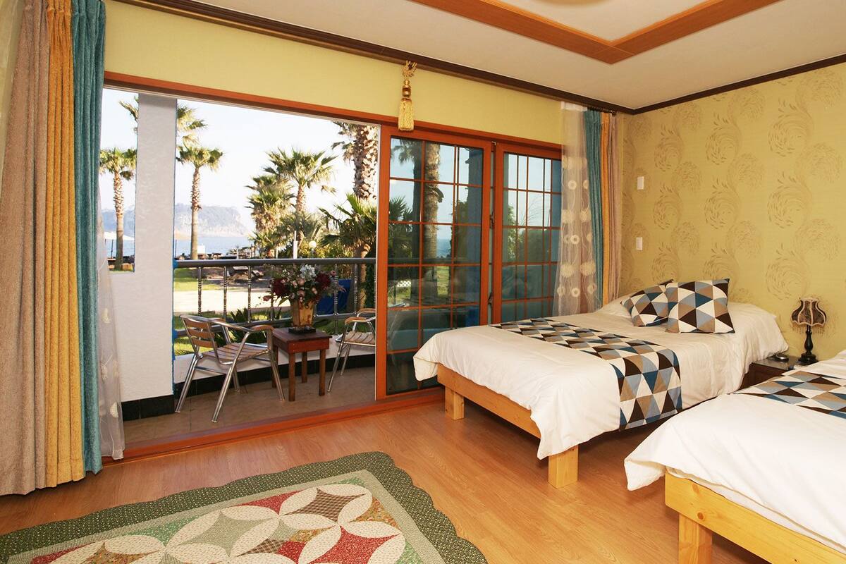 A bedroom with two beds, a balcony with a table and chairs, and a view of palm trees.