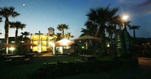 Et resort med udendørs siddepladser, palmer og en bygning med oplyste vinduer.