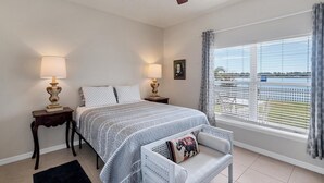 Ein Schlafzimmer mit einem Bett, zwei Nachttischen mit Lampen, einem Deckenventilator, einem Fenster mit Blick auf ein Gewässer und einem Sessel mit Kissen.