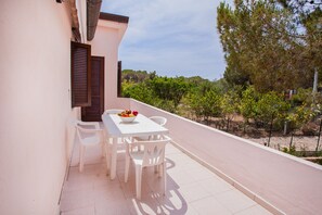 A balcony with a white table and chairs, a bowl of fruit, and a view of trees and a fence.