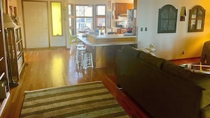 A spacious living room with a dark green sofa, a wooden coffee table, and a striped rug. A kitchen with wooden cabinets and a refrigerator is visible in the background.
