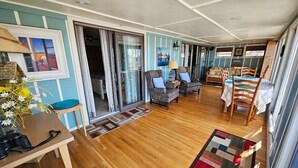 A spacious living area with a wooden floor, a sofa set, and a dining table with chairs. There is a large window providing natural light and a view of the outdoors.