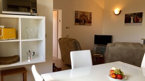 Un salon avec une table blanche, un fauteuil marron, un canapé beige, un téléviseur et un tableau accroché au mur.