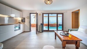 Una cocina moderna con armarios blancos, una encimera de madera y un horno. Hay un comedor con una mesa y sillas de madera. Un amplio ventanal corredera da a una zona de piscina con vistas al océano.