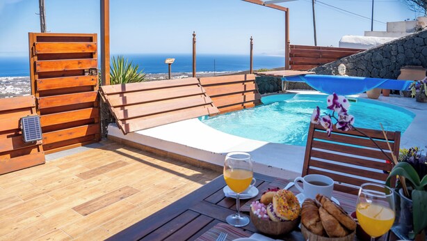 Una terraza de madera con piscina, mesa de desayuno con bebidas y pasteles, y vista al mar.