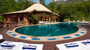 A pool area with a wooden deck, lounge chairs, and a thatched-roof building. - The Rangers Reserve - Corbett (Nainital)