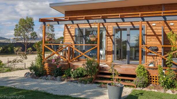 A modern wooden cabin with large windows, a covered porch, and a garden area with flowers and plants. - Harrison Grove (Forcett)