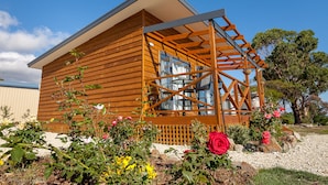 A wooden cabin with a sloped roof, a wooden deck, and a garden with flowers and a large tree. - Harrison Grove (Forcett)