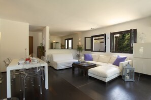 Ein modernes Wohnzimmer mit einem weißen Sofa, einem Couchtisch und einem Essbereich mit weißem Tisch und klaren Stühlen.