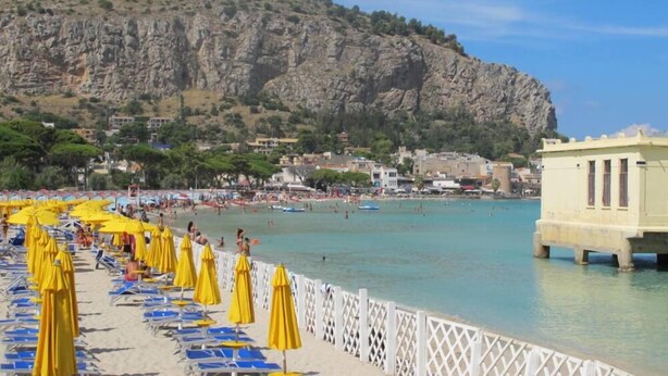 Une plage bordée de rangées de chaises longues et de parasols jaunes, une colline rocheuse en arrière-plan et un ciel bleu dégagé.