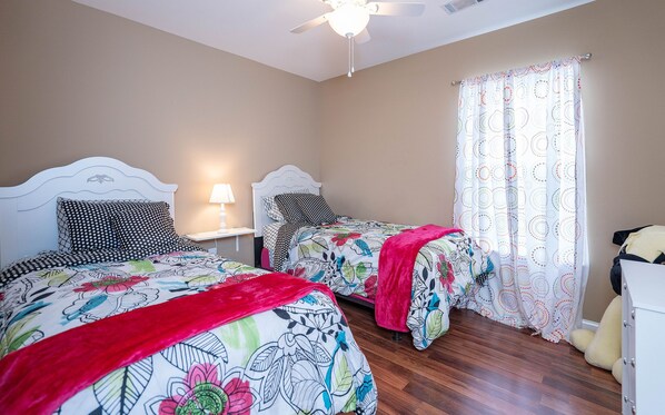 A bedroom with two beds, a ceiling fan, a nightstand with a lamp, and a window with patterned curtains.