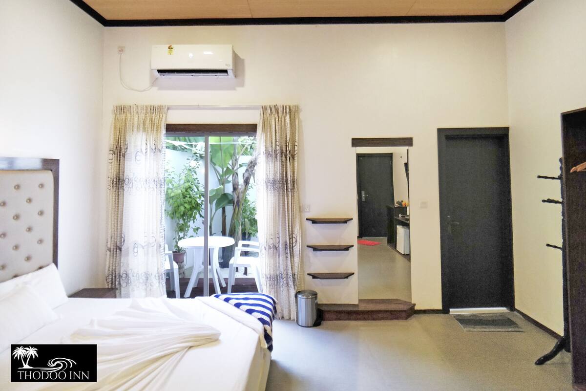 A hotel room with a bed, a small table with chairs, an air conditioning unit, a bathroom with a sink and mirror, and a door leading to another room.
