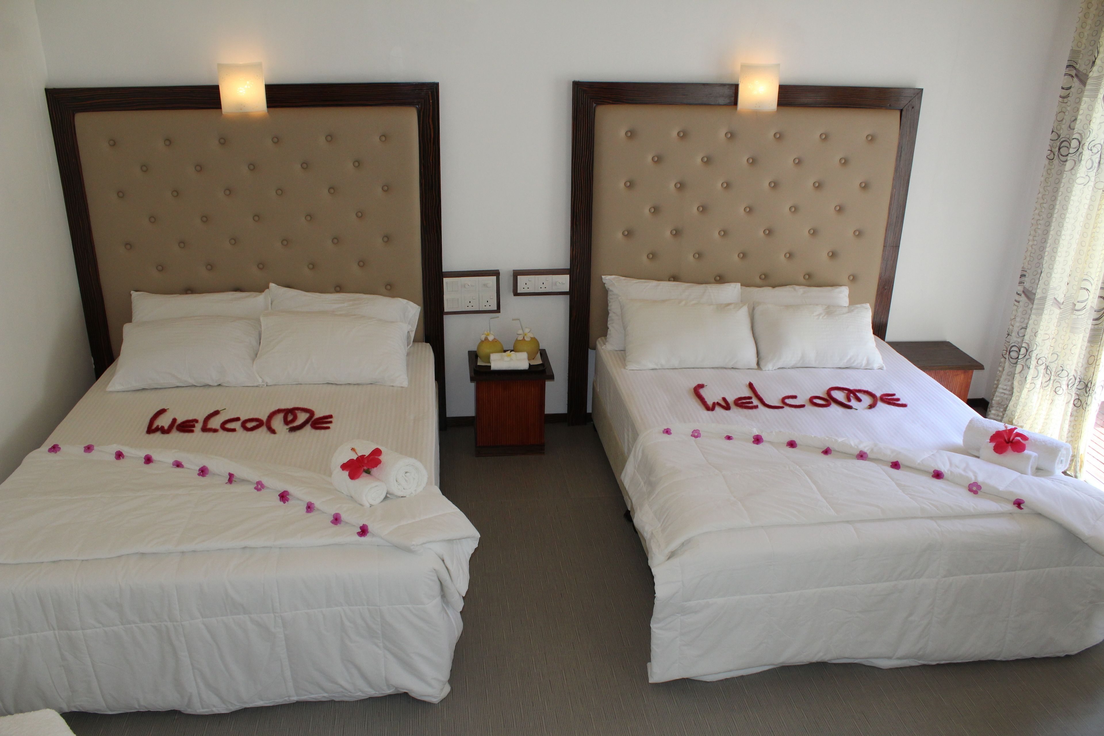 Two twin beds with 'Welcome' written on the bedspread, a small table with a bottle and a glass, and a nightstand with a lamp.