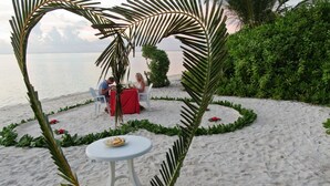 A beachside dining setup with a white table and chairs, surrounded by greenery and palm fronds, overlooking the sea. - Thoddoo Inn (Thoddoo)