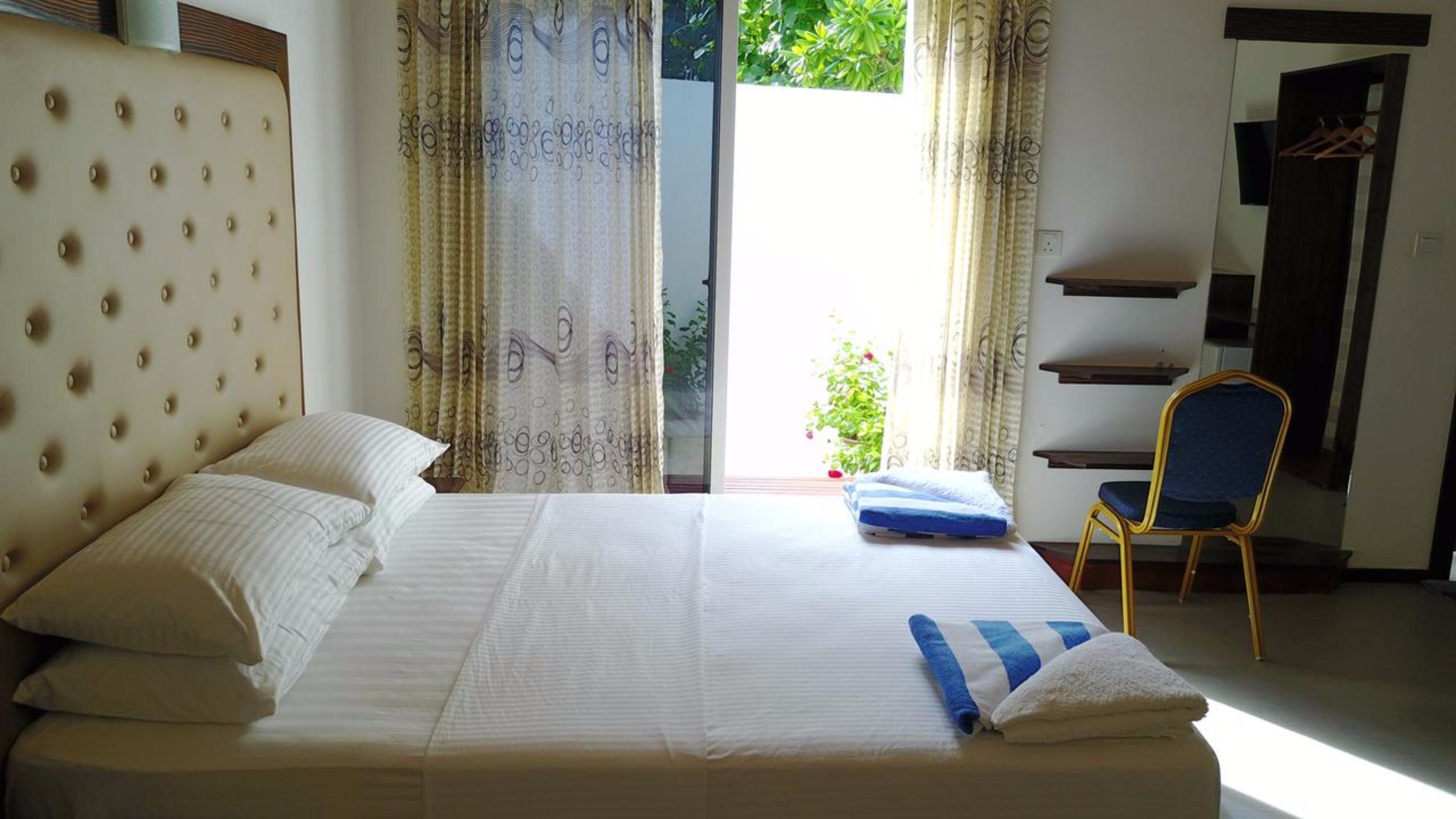 A neatly made bed with white linens and a blue striped pillow, a chair with a yellow frame, and a view of greenery through the window.