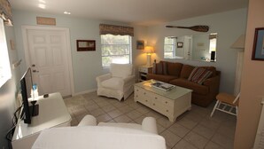 A room with a white door, a white armchair, a brown sofa, a glass coffee table, a white dresser, and a mirror on the wall. - 1st Floor 2 Bed 1 bath home, short walk to beach, 1 step to door, North end LBK (Longboat Key)