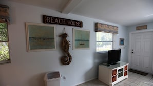 A room with a television on a cabinet, framed paintings, a 'Beach House' sign, and a door. - 1st Floor 2 Bed 1 bath home, short walk to beach, 1 step to door, North end LBK (Longboat Key)
