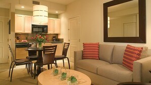 Un salón con un sofá beige, una mesa redonda con objetos decorativos y una zona de comedor con sillas y una cocina al fondo.